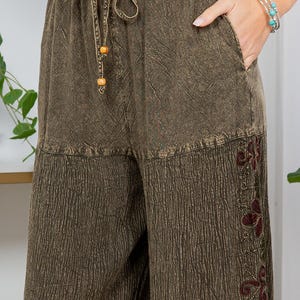 May include: Olive green wide-leg trousers with a drawstring waist and pockets. The trousers feature a textured fabric and embroidered floral details on the lower leg. The hemline has a colourful floral print.