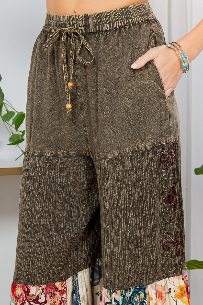 May include: Olive green wide-leg trousers with a drawstring waist and pockets. The trousers feature a textured fabric and embroidered floral details on the lower leg. The hemline has a colourful floral print.