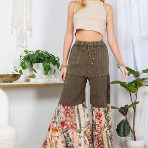 May include: A woman in a beige crop top and olive green wide-leg trousers with floral patchwork and a straw hat. The trousers have a drawstring waist and lace trim. The outfit has a casual, bohemian style.