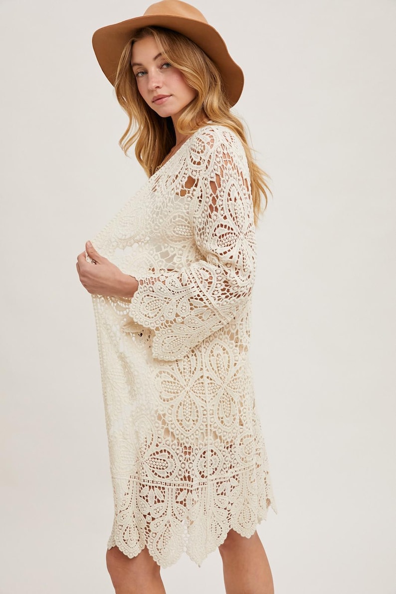 May include: Cream-colored, long-sleeved crochet cardigan with a scalloped hem. The open-front design features intricate floral and geometric patterns. A wide-brimmed brown hat completes the look. This bohemian-style garment is perfect for layering.