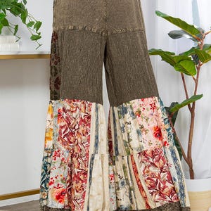 May include: Wide-leg trousers with a patchwork design. The top section is a textured olive green, while the lower part features floral and striped patterns in various colours. The hemline is finished with a brown lace trim.