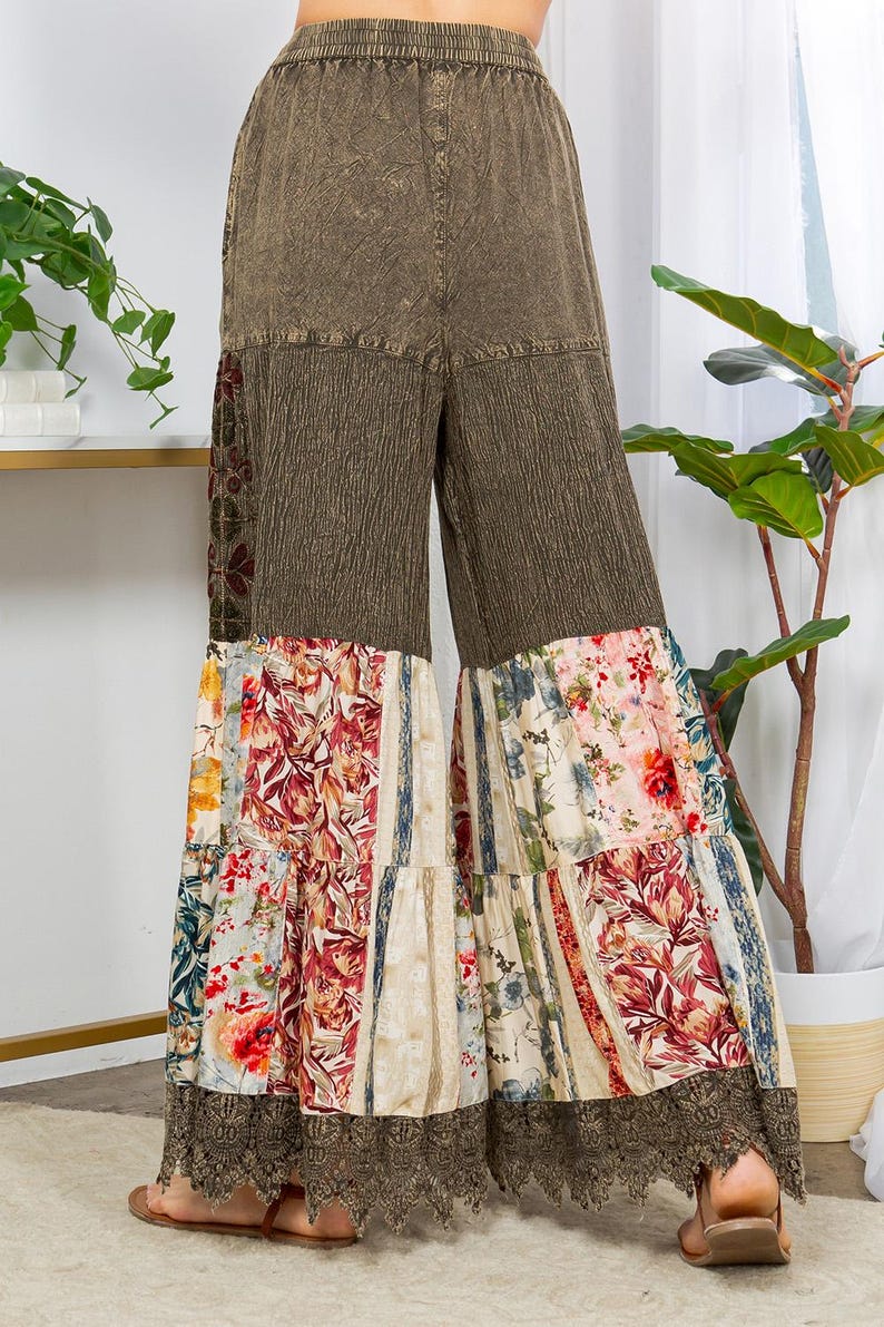May include: Wide-leg trousers with a patchwork design. The top section is a textured olive green, while the lower part features floral and striped patterns in various colours. The hemline is finished with a brown lace trim.