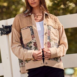 May include: A tan corduroy jacket with floral tapestry pockets and quilted shoulder panels. The jacket has a button-up front and is worn with a white top and a tan hat. The background is a blurred outdoor scene.