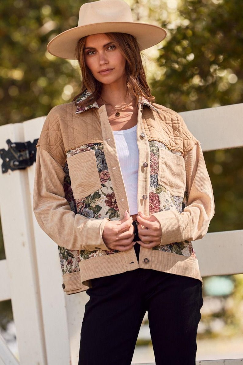 May include: A tan corduroy jacket with floral tapestry pockets and quilted shoulder panels. The jacket has a button-up front and is worn with a white top and a tan hat. The background is a blurred outdoor scene.