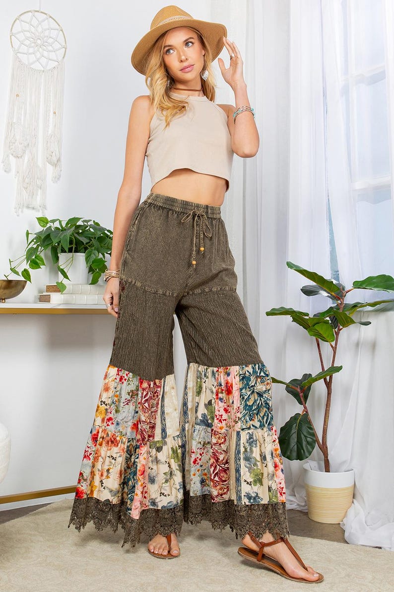 May include: A woman wearing a beige crop top, olive green palazzo trousers with floral patchwork, and a straw hat. The trousers have a drawstring waist and lace trim. The outfit is paired with sandals. A dreamcatcher hangs in the background.