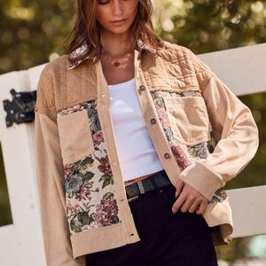 May include: Tan corduroy jacket with floral tapestry accents. The jacket features a quilted shoulder panel, button-up closure, and two front pockets. A beige fedora hat completes the look. The jacket is paired with a white top and black jeans.