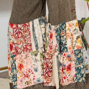 May include: Wide-leg trousers with a patchwork floral design. The upper section is textured olive green, and the lower section features panels of floral prints in red, pink, blue, and cream. The hem is trimmed with brown lace.
