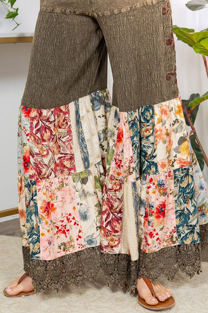 May include: Wide-leg trousers with a patchwork floral design. The upper section is textured olive green, and the lower section features panels of floral prints in red, pink, blue, and cream. The hem is trimmed with brown lace.