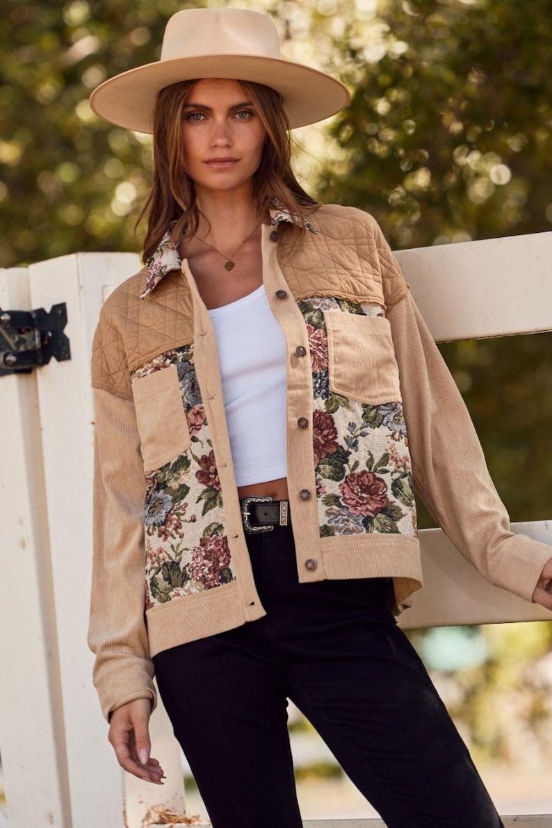 May include: A tan corduroy jacket with floral tapestry panels and quilted shoulder accents. The jacket has a button-up front, two front pockets, and a collared neckline. The model is wearing a white top, black pants, and a tan hat.