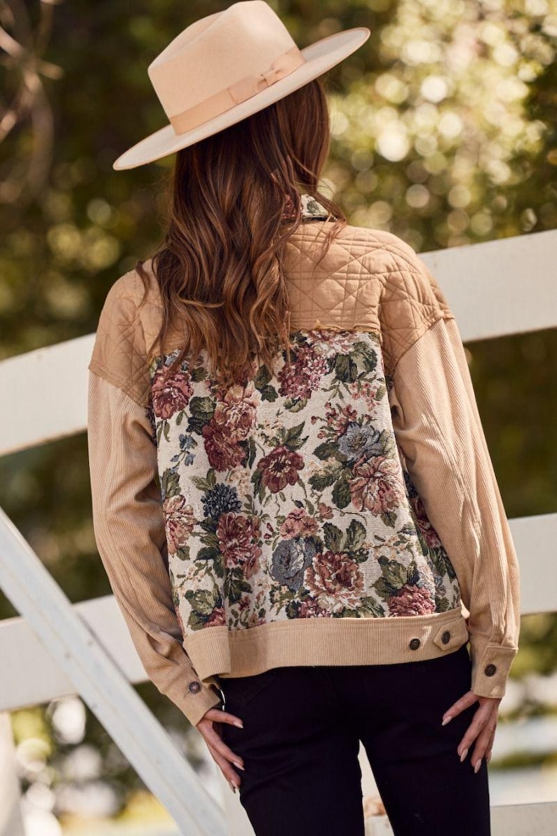 May include: Tan corduroy jacket with a floral tapestry back panel. The jacket features a quilted upper back, buttoned cuffs, and a neutral-toned color palette. A beige hat is worn.