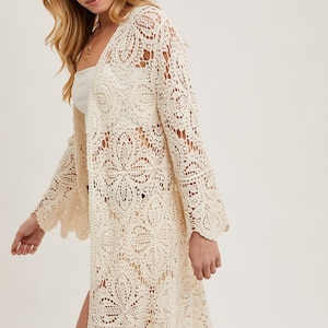 May include: Cream-colored, open-front crochet cardigan with bell sleeves. The long, lightweight garment features an intricate floral lace pattern and a scalloped hemline. A brown hat is also visible.