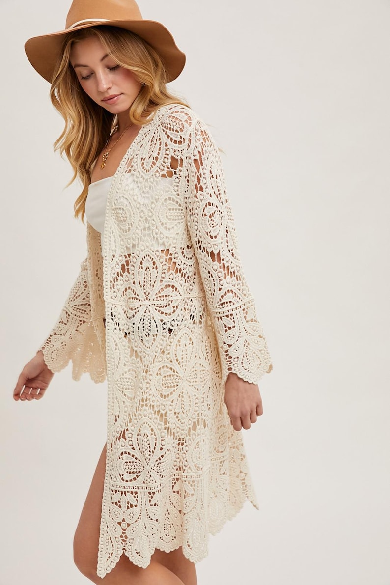 May include: Cream-colored, open-front crochet cardigan with bell sleeves. The long, lightweight garment features an intricate floral lace pattern and a scalloped hemline. A brown hat is also visible.