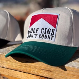 May include: A beige baseball cap with a green brim. The cap features a patch with a red and white design and the text "GOLF CIGS DON'T COUNT" in black letters. The cap is sitting on a wooden surface.