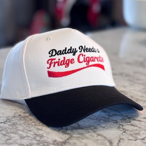 Könnte beinhalten: Eine weiße Baseballkappe mit schwarzem Schirm. Auf der Kappe steht der Text "Daddy Needs a Fridge Cigarette" in schwarzer und roter Schrift. Der Hut ist aus robustem Material gefertigt und für den täglichen Gebrauch geeignet.