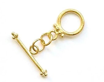 May include: A gold-colored toggle clasp, featuring a circular ring and a straight bar with decorative ends. The clasp is connected by small, linked rings, and is set against a white background. This jewelry finding is ideal for necklaces and bracelets.