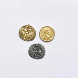 May include: Three small, round pendants and a penny on a white background. Two pendants are gold-toned, one with a starburst design. The third pendant is silver-toned, also with a starburst design. The penny is gold-toned with a portrait.