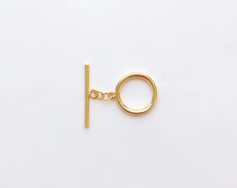 May include: A gold-colored toggle clasp, featuring a straight bar and a circular ring connected by a small chain. The clasp is set against a plain white background, highlighting its simple design and metallic finish.