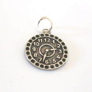 May include: A silver-colored clock charm with a circular face. The clock face features black dots around the edge, with numbers 1 through 12. The hands of the clock are pointing to approximately 2:40. A small loop is at the top for attaching to a chain.