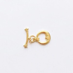 May include: A gold-colored toggle clasp featuring a crescent moon with a smiling face and a star, along with a bar with two stars. The clasp is designed for jewelry making and has a celestial theme.