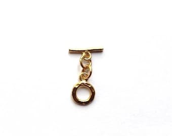 May include: A gold-colored toggle clasp, a jewelry component, is displayed against a white background. The clasp consists of a bar and a ring connected by a series of small, linked rings. The ring has a hammered texture.