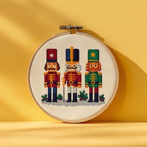 May include: A cross-stitch embroidery hoop featuring three pixelated nutcracker soldiers. The soldiers wear red, blue, and green uniforms with black boots. The embroidery is set against a cream-colored fabric and framed by a wooden hoop.