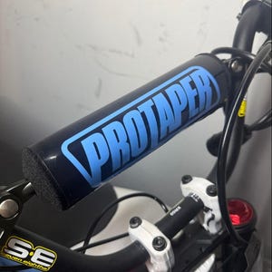 May include: Black handlebar pad with the word "PROTAPER" in light blue. The pad is on a black handlebar with white and black components. The image also shows the "SE" logo and other bike parts.