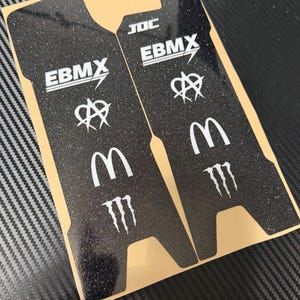 May include: Two black, glitter-textured stickers with white logos. The stickers feature the text "EBMX", "JDC", and the McDonald's and Monster Energy logos. The stickers are on a tan backing and are likely for a skateboard or BMX bike.