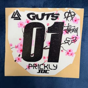May include: White sticker with black glitter number "01" and the word "GUTS" at the top. Pink cherry blossom branches and the word "PRICKLY" are also visible. Various logos and symbols are also present.