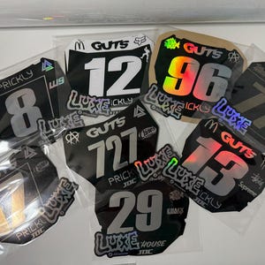 May include: A collection of motocross number plates in clear plastic sleeves. The plates are black with white or holographic numbers and logos. Various brands and designs are visible, including "GUTS", "LUXE", and "PRICKLY".