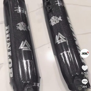 Puede incluir: Dos tubos de horquilla de motocicleta negros con logotipos y texto plateados. Los tubos tienen un acabado brillante y presentan la marca "Dunlop", un logotipo estilizado y el logotipo de la bebida energética "Monster".