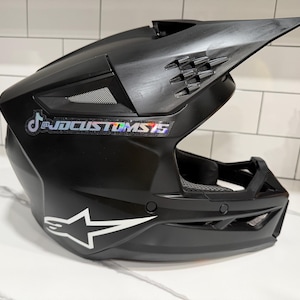 May include: Black motocross helmet with a visor and vents. Features a white star logo and a sticker with the text "@JDCUSTOMS15". The helmet is designed for protection and performance.