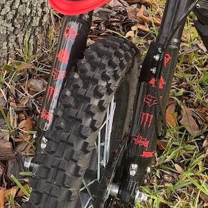 Puede incluir: Primer plano del neumático delantero y la suspensión de una moto de cross negra y roja. El neumático tiene un dibujo de tacos. La suspensión tiene gráficos y logotipos rojos. La bicicleta está sobre hierba cerca de un árbol.