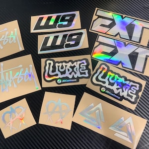 Op de afbeelding: Een verzameling holografische stickers met diverse ontwerpen en tekst. De stickers bevatten het "Stussy"-logo, het woord "LUXE" met de tekst "JDcustoms", het woord "EXT", de letters "W9" en abstracte symbolen. De stickers liggen op een bruinachtige achtergrond.