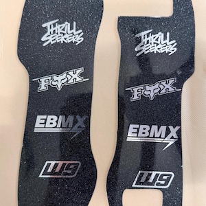 Peut inclure: Deux coussinets de préhension noirs et texturés avec des logos blancs. Les logos incluent "Thrill Seekers", "FOX", "EBMX" et "119". Un coussinet a un dessus incurvé avec "JDC" imprimé dessus.