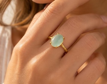 May include: A gold ring featuring a smooth, oval-shaped, pale mint green cabochon gemstone set in a four-prong setting. The band of the ring has a textured, slightly irregular surface, providing a distinct contrast to the polished stone.