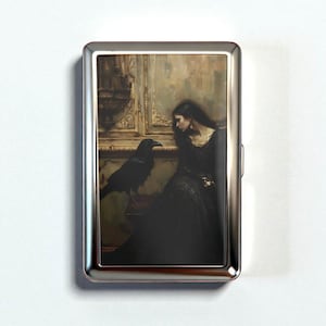 May include: A silver cigarette case featuring a gothic painting of a woman in a black dress with a raven. The artwork has a vintage aesthetic with muted colors and a dramatic composition. The case has a shiny, metallic finish.