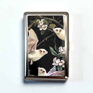 May include: A silver metal cigarette case featuring a detailed illustration of three white bats with outstretched wings, set against a dark background with white flowers and branches. The bats are the focal point of the design.