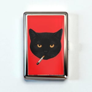 May include: A silver cigarette case with a red background featuring a black cat with yellow eyes and a cigarette in its mouth. The case has a rectangular shape and a metallic finish.