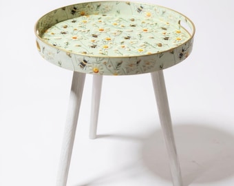 Puede incluir: Una mesa auxiliar redonda con una tapa verde claro con un estampado floral y de abejas. La mesa tiene un borde dorado y tres patas de madera blancas. La mesa tiene aproximadamente 50 cm de diámetro y 50 cm de alto.