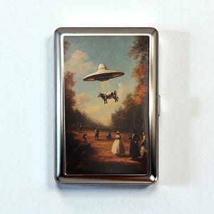 May include: A silver metal cigarette case featuring a surreal painting of a cow being abducted by a UFO. The artwork depicts a rural scene with figures observing the event under a cloudy sky and autumn trees.