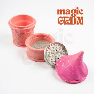 May include: A disassembled pink grinder with a cone-shaped top, a mesh screen, and a silver grinding plate. The grinder is made of pink silicone and has the words "magic GRDN" in orange.