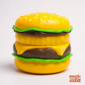 May include: A yellow and brown burger-shaped container with sesame seeds on top. The burger has green lettuce, yellow cheese, and a brown patty. The container is designed to look like a hamburger.