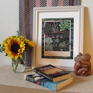 Pode incluir: Uma impressão emoldurada de uma ilustração botânica que retrata uma variedade de plantas e vegetais em um jardim. A impressão está em uma moldura branca e é exibida em uma mesa com um vaso de girassóis, dois livros e uma escultura de madeira.