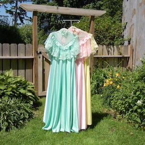 May include: Three pastel-colored vintage gowns hang on a wooden rack outdoors. The gowns are mint green, pink, and yellow, with lace detailing on the bodice and sleeves. The gowns are long and flowing, with a satin-like sheen. The background includes a wooden fence and lush greenery.