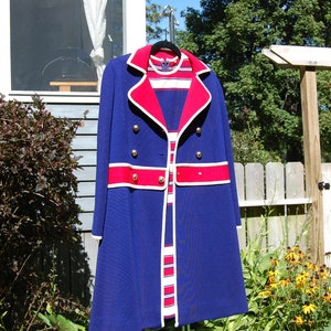 May include: A vintage blue coat with red and white accents, featuring gold buttons. The coat is hanging outside on a sunny day. The coat has a matching dress underneath.