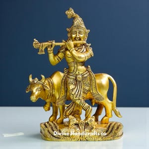 May include: A golden brass sculpture of a deity playing a flute while riding a bull. The sculpture has intricate details and is set on a decorative base. The text "Divine Handicrafts.co" is visible on the base.