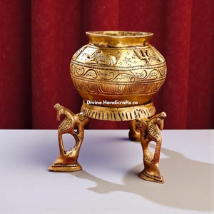 May include: A gold-colored, ornate metal pot with intricate carvings and a wide rim. The pot rests on three decorative legs, each featuring a bird-like design. The text "Divine Handicrafts co" is visible on the pot.