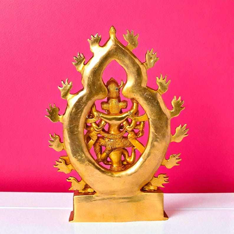 Puede incluir: Una estatua dorada de una deidad con m&uacute;ltiples brazos, ubicada dentro de un marco ornamental en forma de llama. La estatua est&aacute; sobre una base rectangular y el fondo es de color rosa vibrante. Es probable que sea un art&iacute;culo religioso o decorativo.