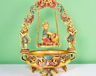 Può includere: Una scultura decorativa dorata con una figura seduta su un'altalena, adornata con pietre colorate intarsiate. Due figure affiancano l'altalena e la base ha la forma di una ciotola. Il testo "Divine Handicrafts co" è visibile.