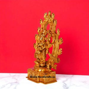 Puede incluir: Una estatua dorada de una deidad con detalles intrincados, sobre un fondo rojo. La estatua presenta m&uacute;ltiples brazos y est&aacute; adornada con adornos ornamentados. La base de la estatua tiene el texto "Divine Handicrafts co".